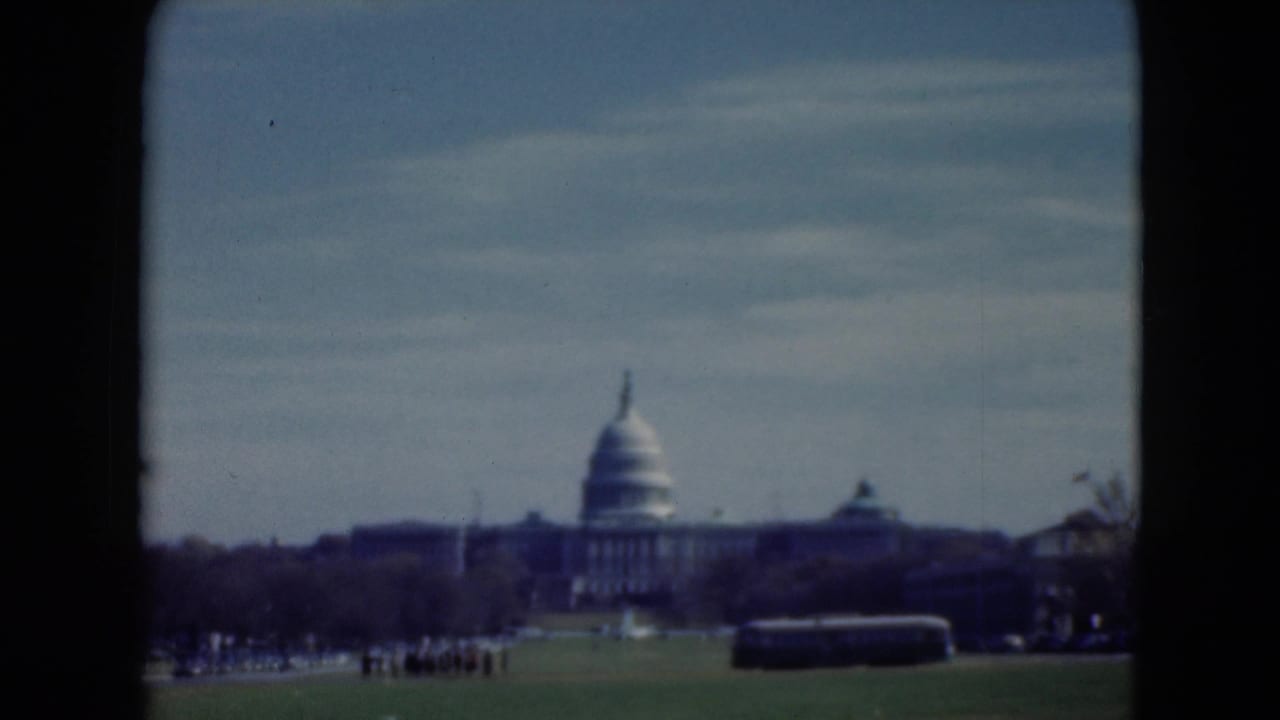 Distant 1975 home-movie frame of the U.S. Capitol area, with a dark bus crossing the foreground and visitors scattered on the grass.