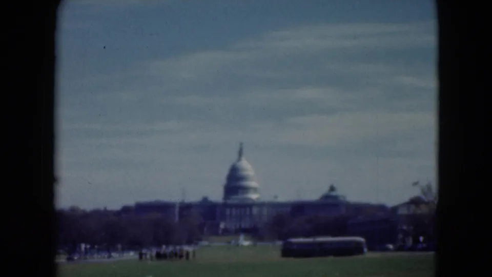 Distant 1975 home-movie frame of the U.S. Capitol area, with a dark bus crossing the foreground and visitors scattered on the grass.