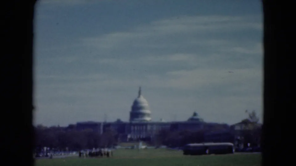 1970s 8mm frame showing a far view of the Capitol dome with a dark bus and visitor groups in the lower foreground.