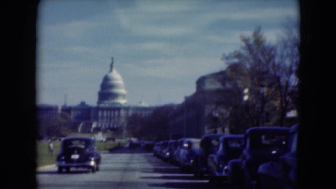 1975 8mm frame of the U.S. Capitol seen down a Washington, D.C. roadway, with parked cars, a moving dark car, trees, and blue sky.