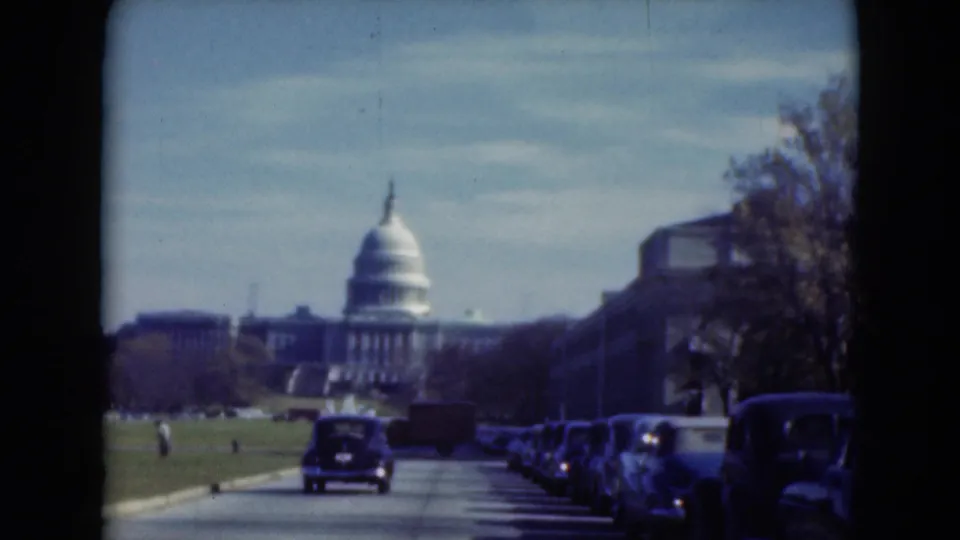 1975 8mm roadside view in Washington, D.C., looking toward the U.S. Capitol with rows of parked cars, traffic, lawn, and bare trees.