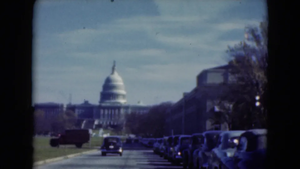 1975 8mm view toward the U.S. Capitol, with parked cars along the curb, a moving car on the road, and a red vehicle crossing the scene.