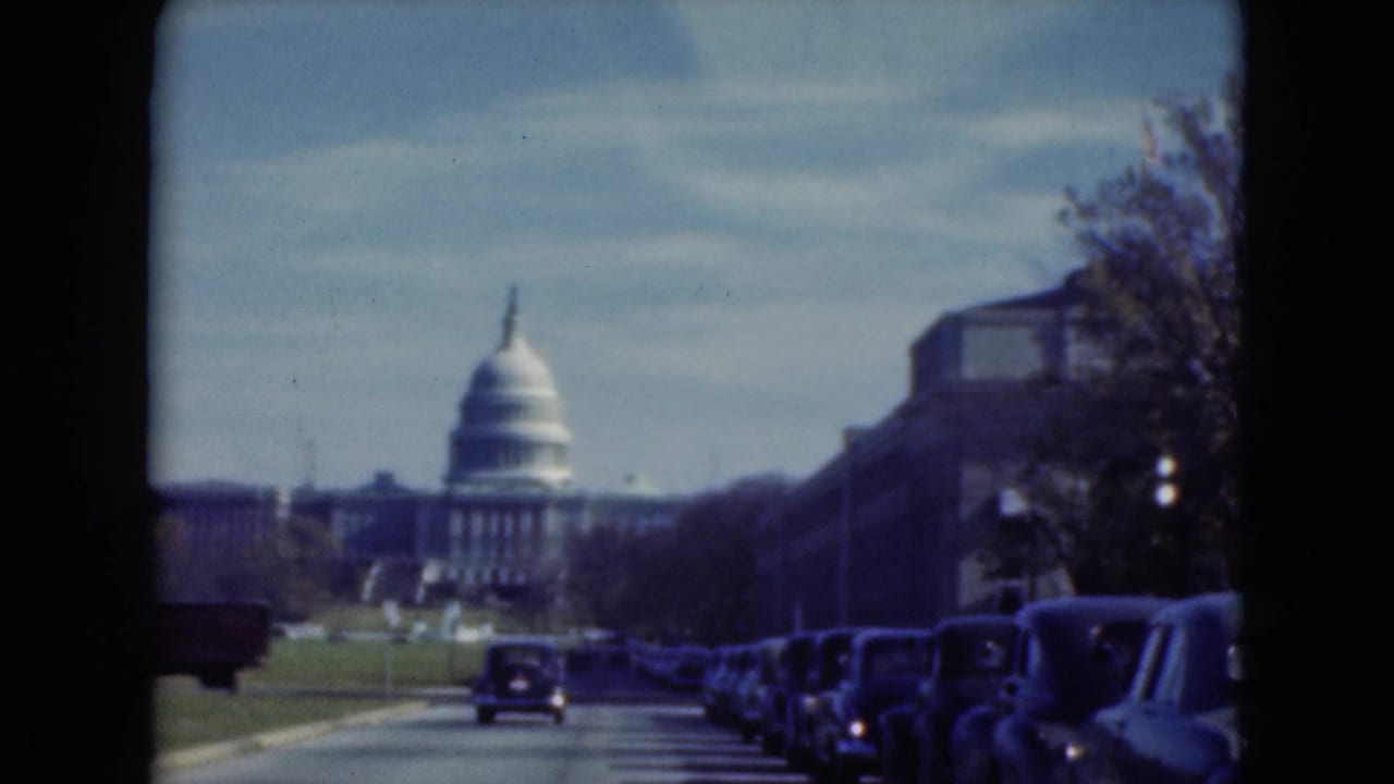 1970s 8mm frame showing a street approach toward a domed civic building, with older cars parked under a bright sky.