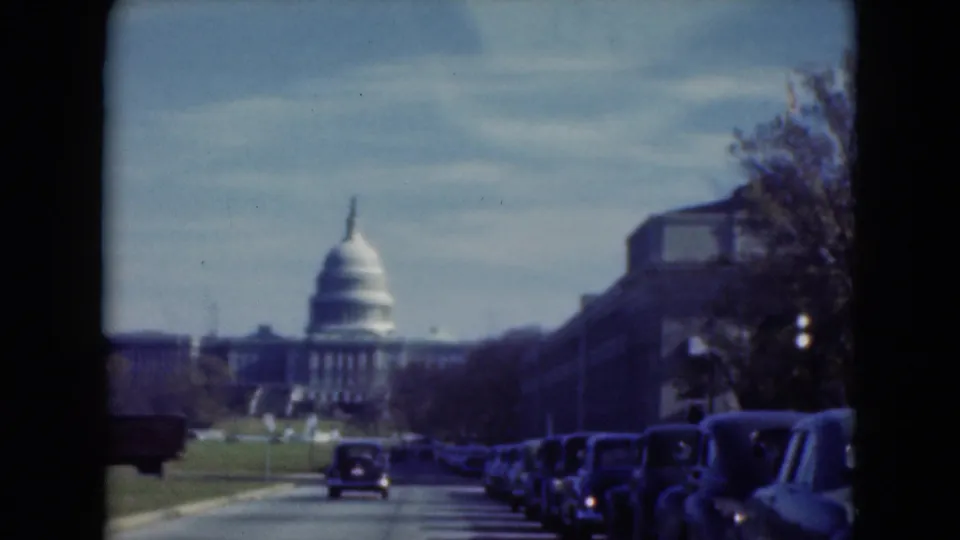 1970s 8mm frame showing a street approach toward a domed civic building, with older cars parked under a bright sky.