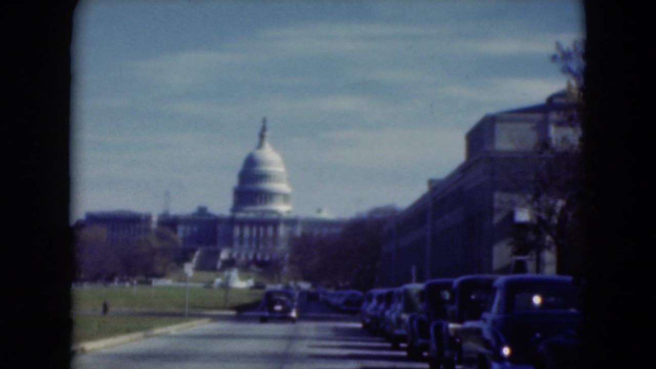 1970s home-movie view down a broad road toward a Capitol-like dome, with parked cars lining the right side.