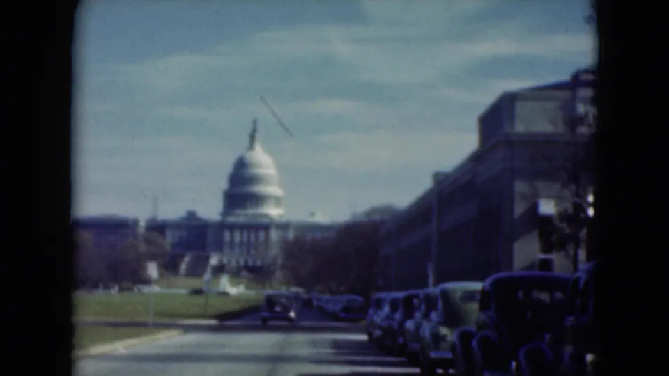 Soft 8mm frame of a road leading toward a domed civic landmark, with 1970s cars and roadside trees visible.