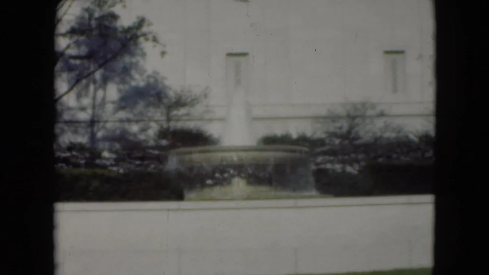 1970s 8mm cutaway of a landscaped fountain with water spray, shrubs, bare trees, and a pale exterior wall.