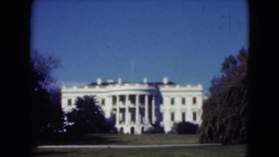 1975 8mm frame showing the White House exterior from a visitor-like vantage, with lawn, trees, blue sky, and soft film focus.