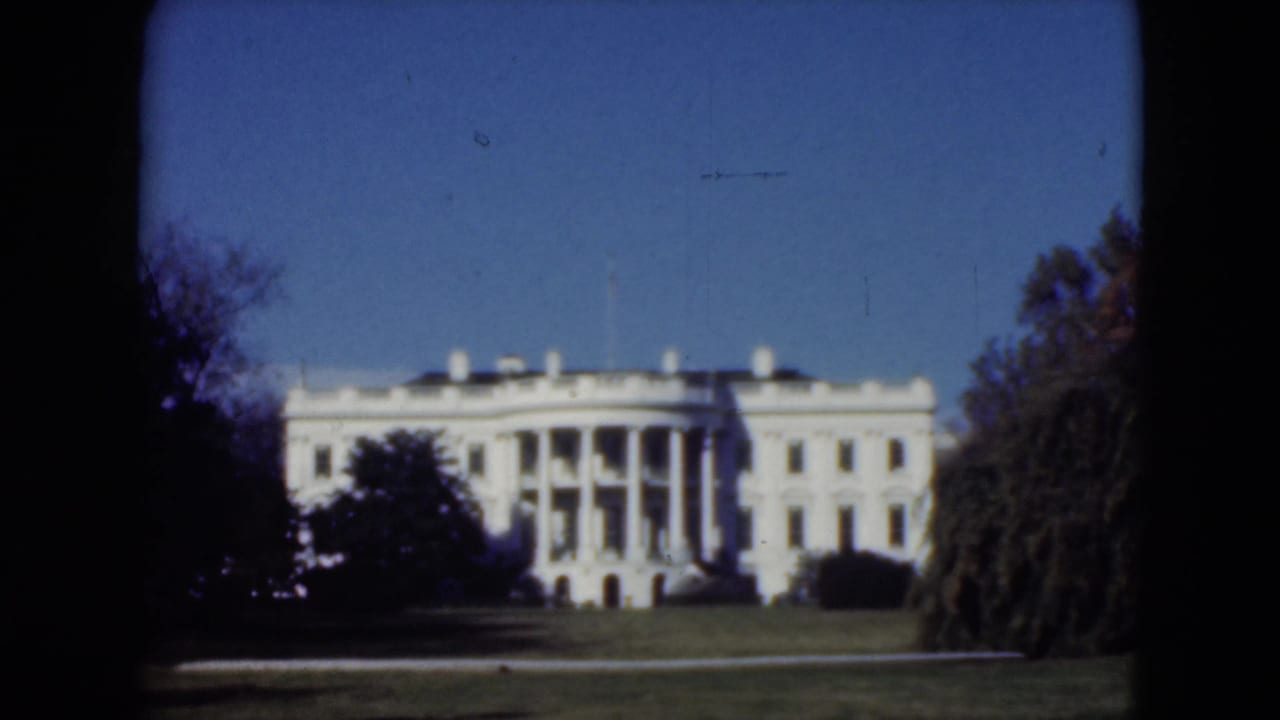 Distant white columned building behind lawn and trees under a blue sky.