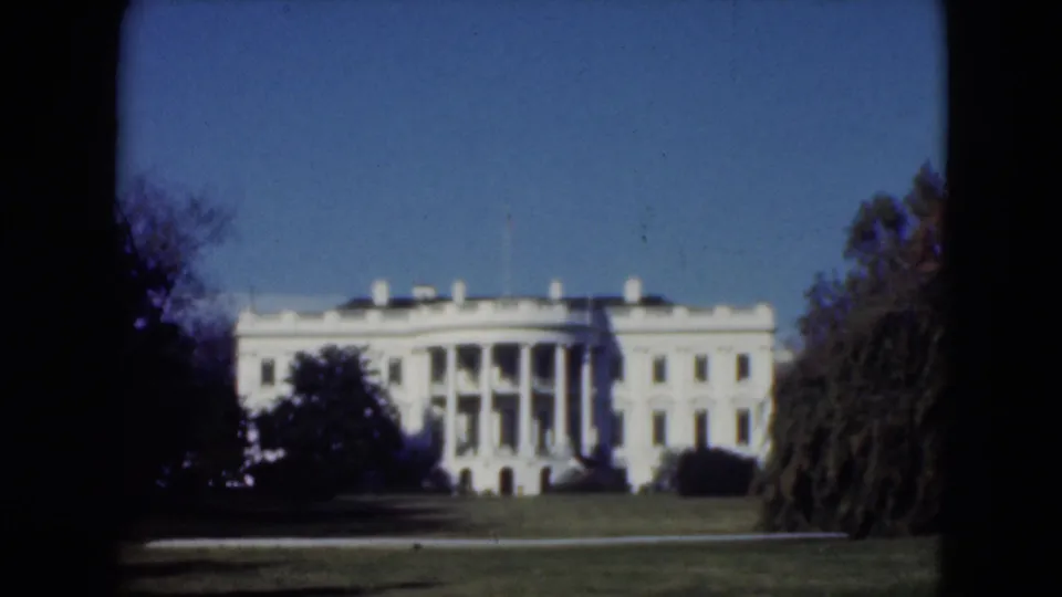 A soft 8mm view of the white monumental building, with lawn in front and trees partly obscuring the facade.