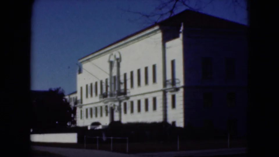 A nearby white building exterior appears from the side, with rows of windows, a lamppost, railings, and deep shadows.