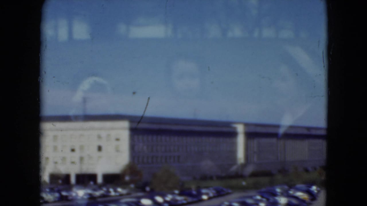 1975 8mm view in Middletown, Pennsylvania of a long institutional building, parked cars, blue sky, and window reflections.