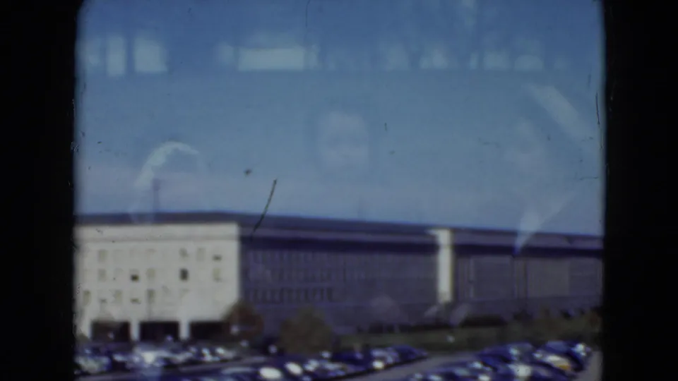 1975 8mm view in Middletown, Pennsylvania of a long institutional building, parked cars, blue sky, and window reflections.