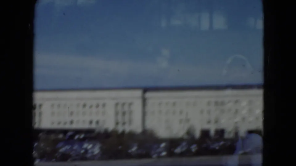 1975 home-movie frame looking past glass at a large Pennsylvania institutional building with cars, window rows, and heavy film borders.