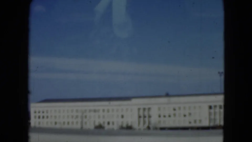 Through the lower pane, the 1975 8mm footage shows a pale institutional facade softened by glass reflections and blue color cast.