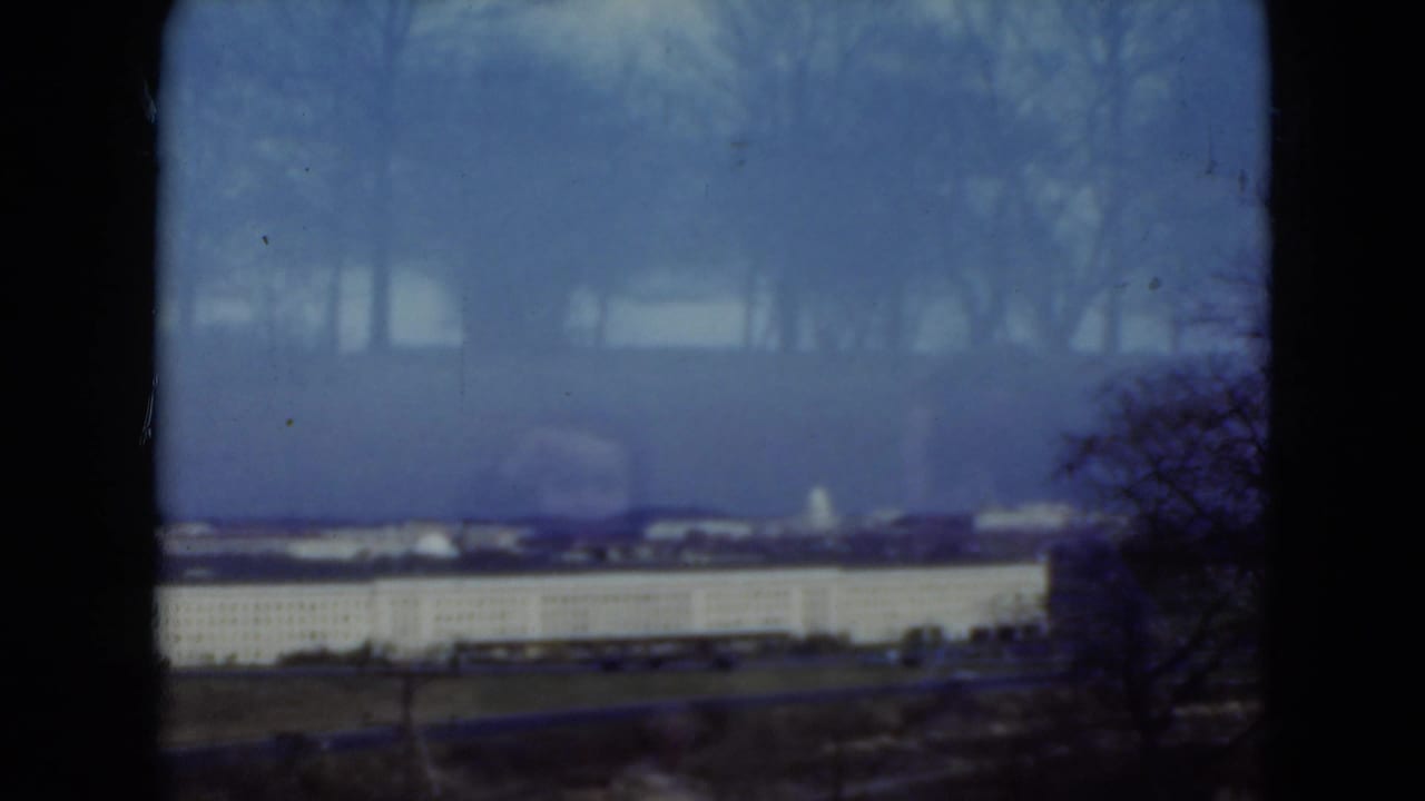 Hazy blue view of a long pale building beyond grass and bare trees, with dark film-frame edges.
