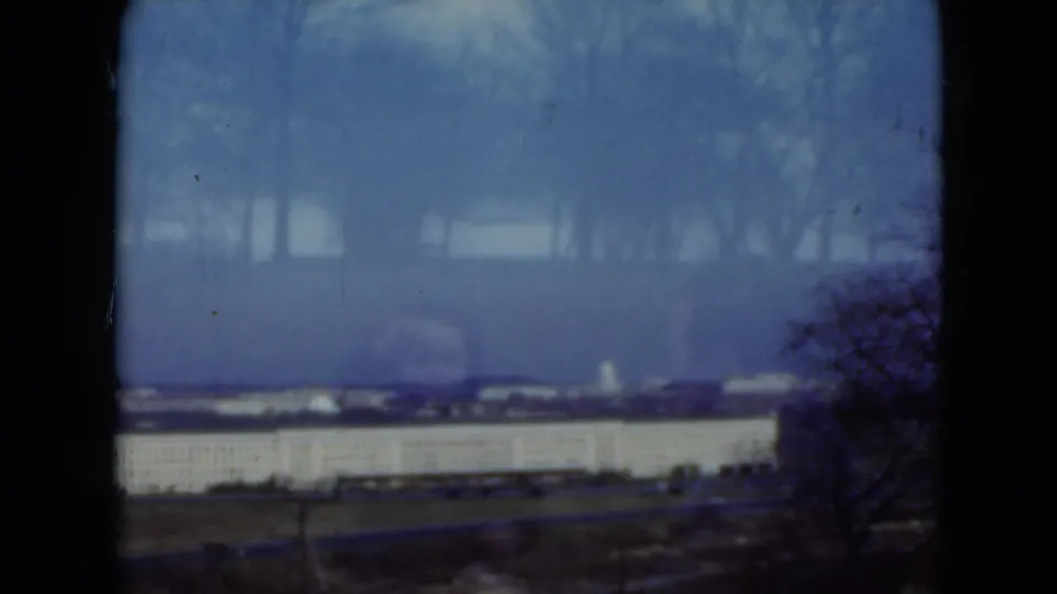 1975 Middletown, Pennsylvania home-movie frame showing a distant pale building through a blue cast and dark gate edges.