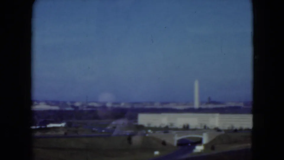 1975 8mm frame showing a moving-vehicle view toward a distant monument and low civic building under a blue sky.