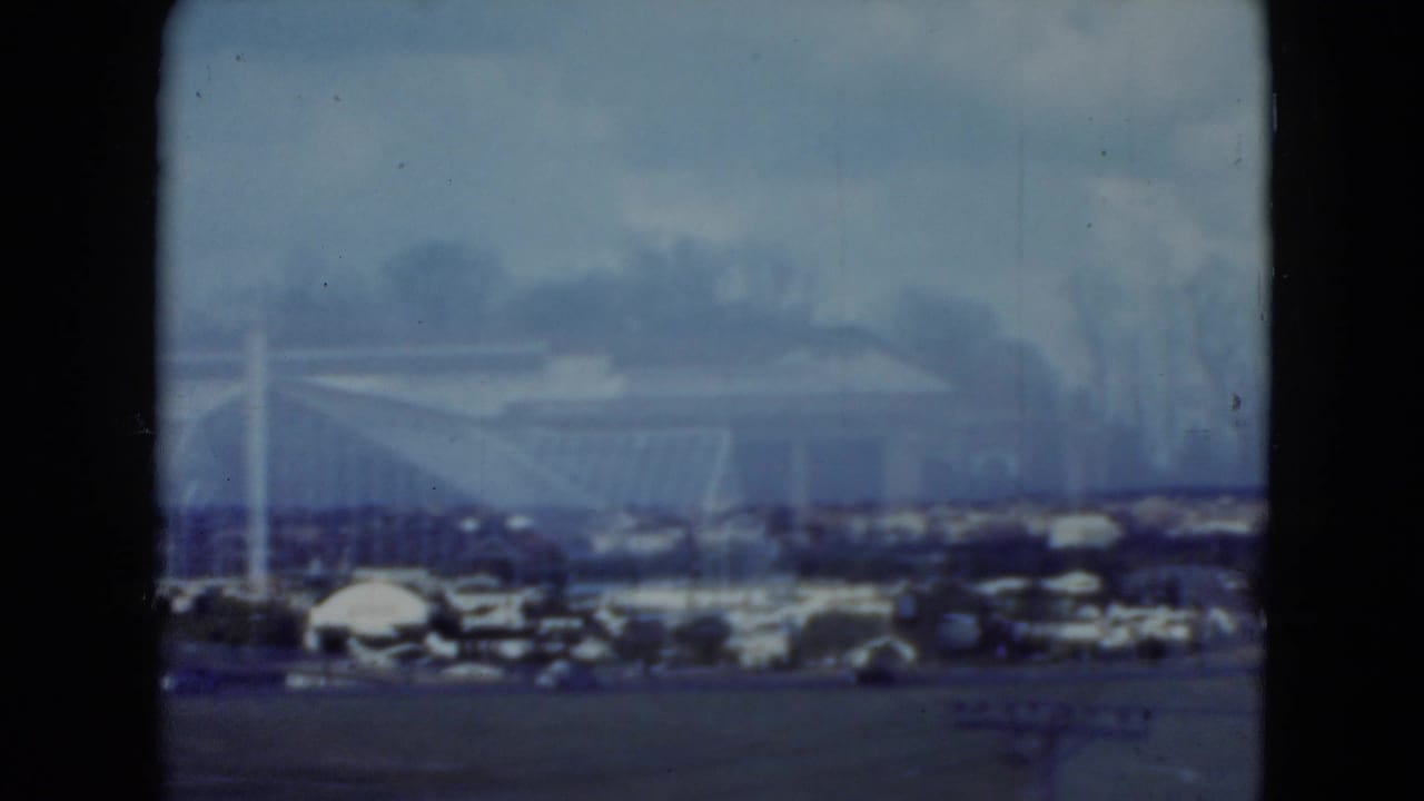 Distant racetrack structure and parked cars framed by dark 8mm film borders.