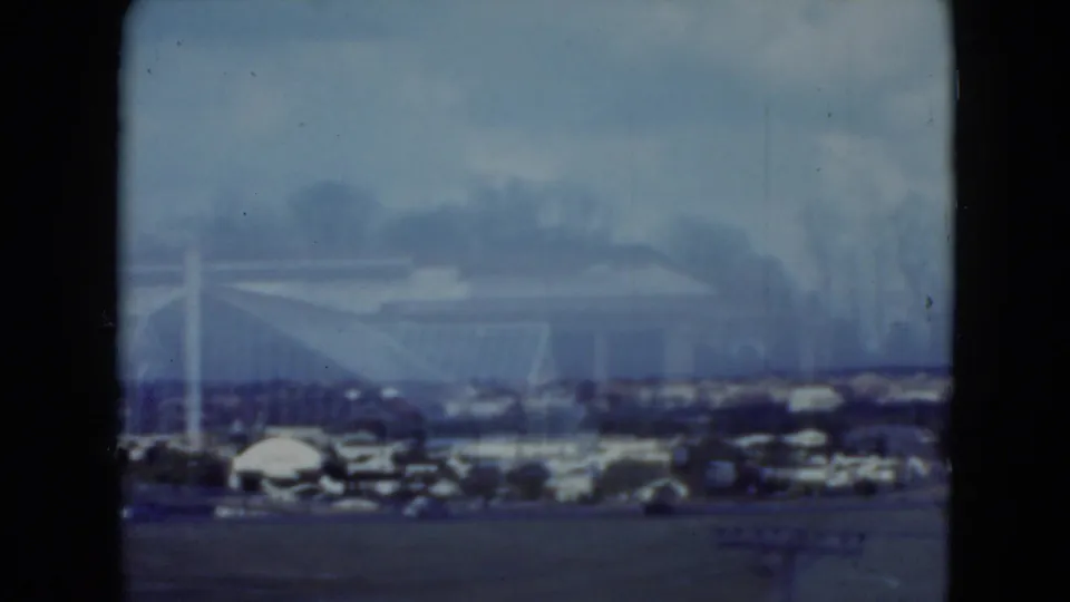 1975 Middletown, Pennsylvania home-movie view of a distant racetrack building, parked cars, and sky through dark film-frame borders.