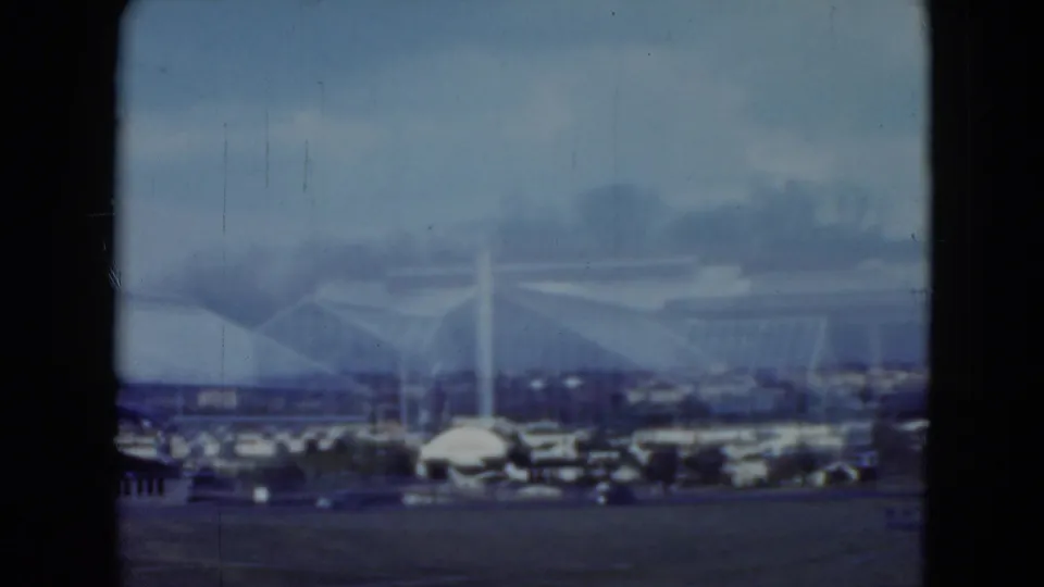 Distant 1975 racetrack or grandstand view in Middletown, with an arched roofline, cars, and dark vertical 8mm frame edge.