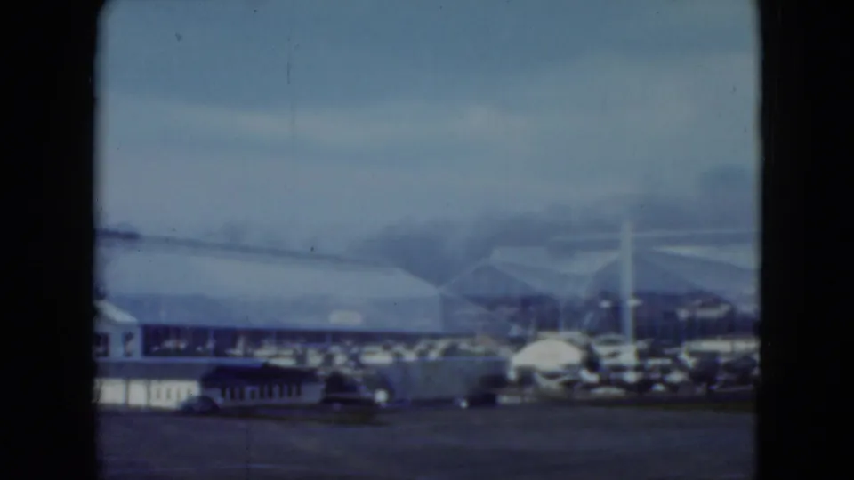 Home-movie frame from Middletown in 1975 showing a distant arched racetrack building, low foreground structure, and parked cars.