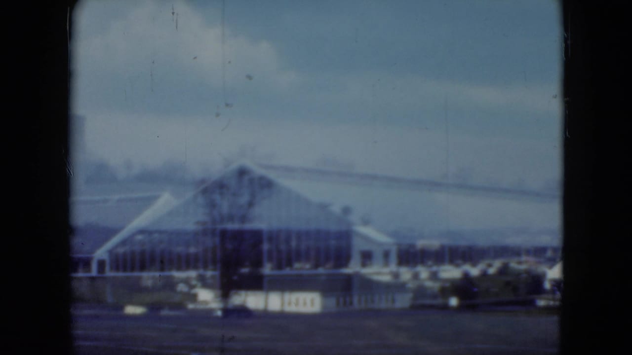 Distant industrial buildings seen through dark 8mm frame borders under a gray sky.