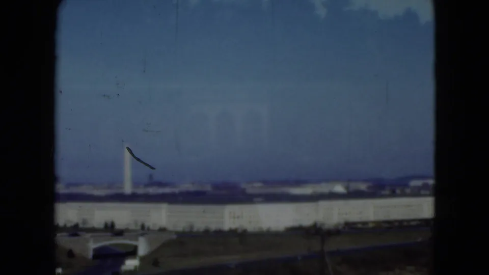 Wide 1975 home-movie view of the Middletown industrial landscape with a tall white tower, low structures, and visible film marks.