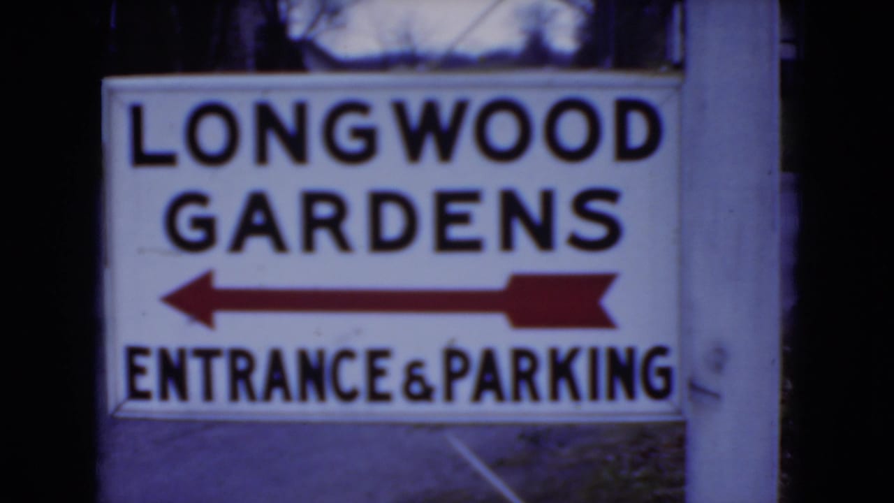1975 8mm frame showing a Longwood Gardens entrance and parking sign on a post in Pennsylvania, with blurred trees behind it.