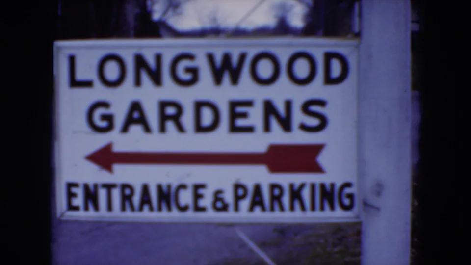 1975 8mm frame showing a Longwood Gardens entrance and parking sign on a post in Pennsylvania, with blurred trees behind it.