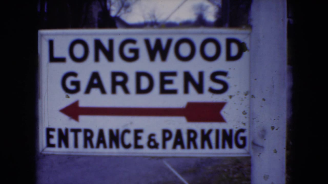 1975 8mm frame of the Longwood Gardens visitor sign, with a red arrow pointing left toward entrance and parking.