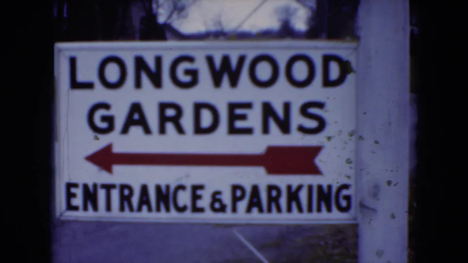 1975 8mm frame of the Longwood Gardens visitor sign, with a red arrow pointing left toward entrance and parking.