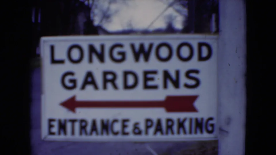 Final 1975 8mm frame showing the Longwood Gardens sign beside a post, with bold lettering and a red left arrow.