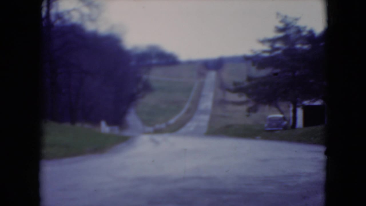 1975 8mm view near the Longwood Gardens approach, showing a paved road climbing through lawns and trees under an overcast sky.