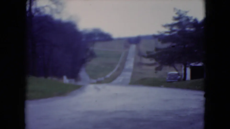 1975 8mm view near the Longwood Gardens approach, showing a paved road climbing through lawns and trees under an overcast sky.