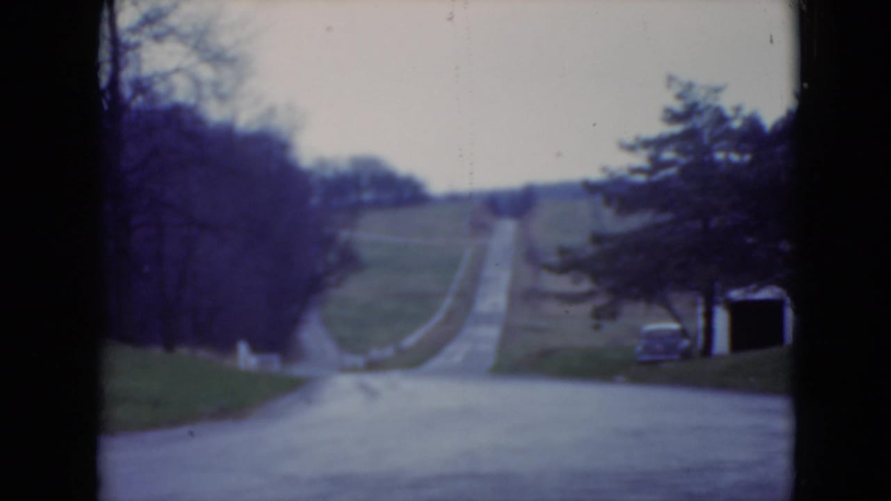 1975 home-movie frame shows the approach road with trees, lawns, and a parked car beside a small roadside structure.