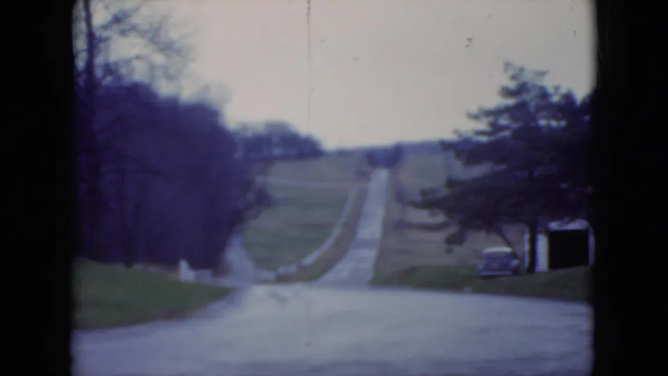 Soft 1975 8mm frame of a Pennsylvania garden approach road, with lawns, bare trees, and a darker evergreen at the right edge.