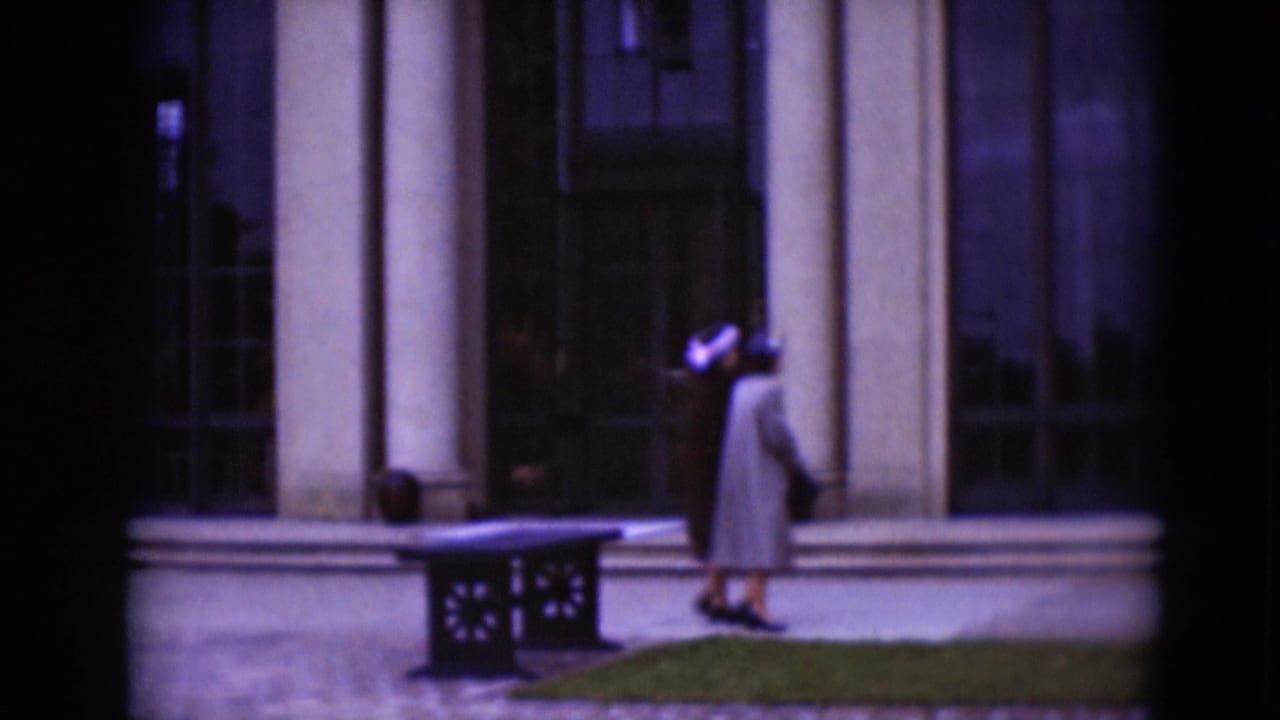 Two women in 1970s attire appear near a columned building exterior with gridded windows, trimmed shrubs, grass, and a low bench.