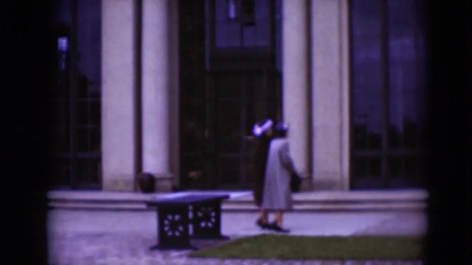 Two women in 1970s attire appear near a columned building exterior with gridded windows, trimmed shrubs, grass, and a low bench.