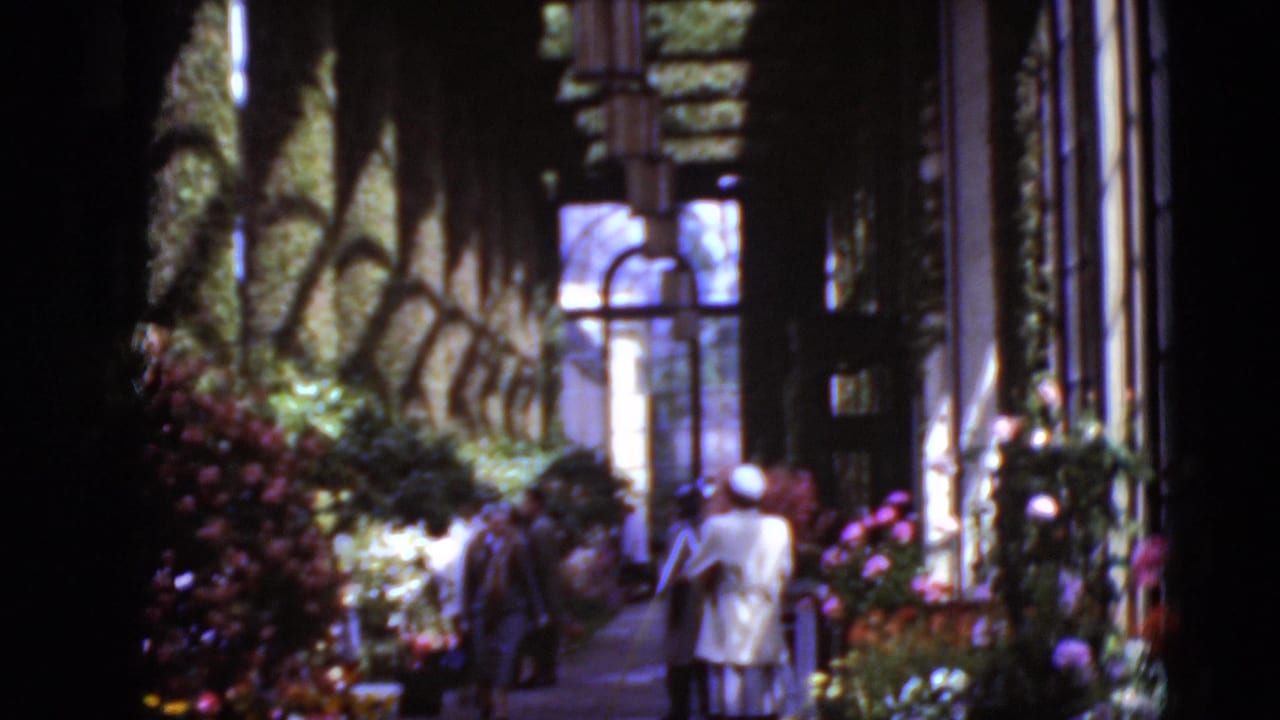 Two women walk through a sun-dappled garden corridor in 1975 Middletown, Pennsylvania, beside flowers, greenery, windows, and a building wall.