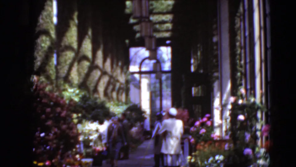 Two women walk through a sun-dappled garden corridor in 1975 Middletown, Pennsylvania, beside flowers, greenery, windows, and a building wall.