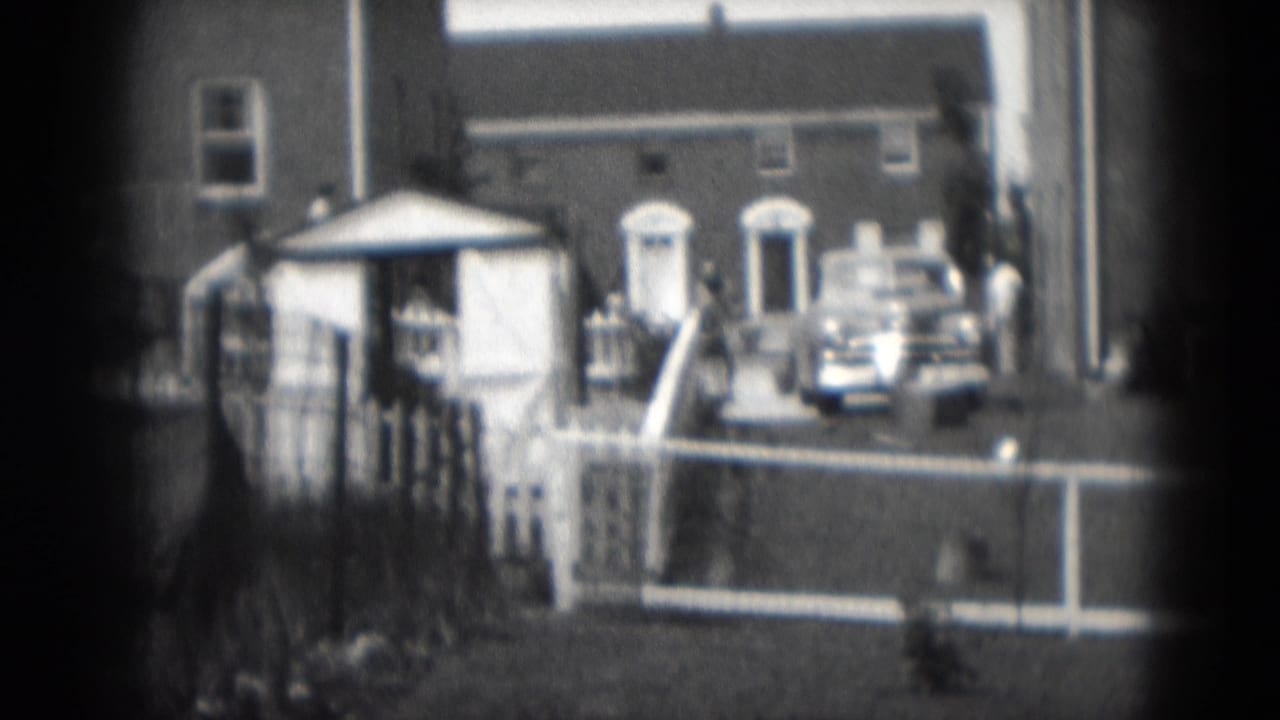 Brick houses, white fence, steps, small outbuilding, and a parked car in a driveway.