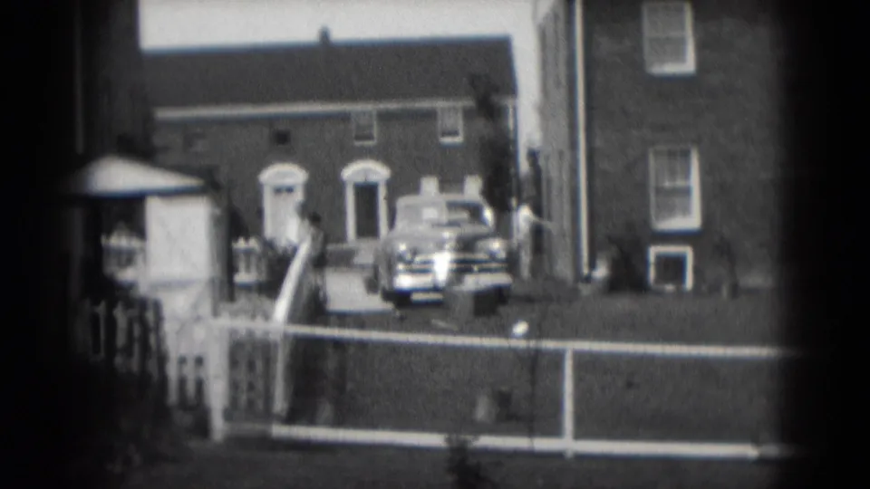 1975 Middletown, Pennsylvania home-movie frame showing brick houses, a white fence, front steps, and an older car parked near a driveway.
