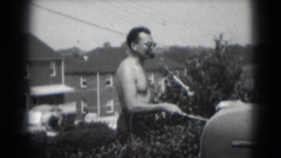 In Middletown, Pennsylvania in 1975, a shirtless man smokes a large cigar while standing by a backyard grill.