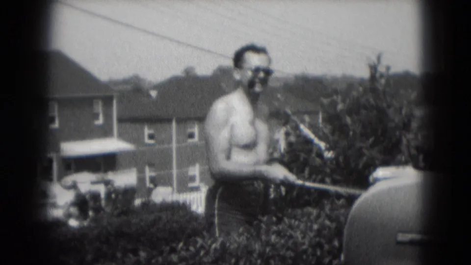 The shirtless man, wearing sunglasses and smoking a cigar, stands near a grill with suburban houses behind him.