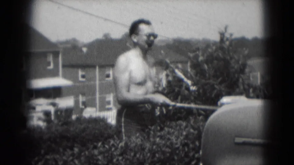 A 1975 home-movie frame shows the man smoking while standing at a backyard grill in suburban Middletown.