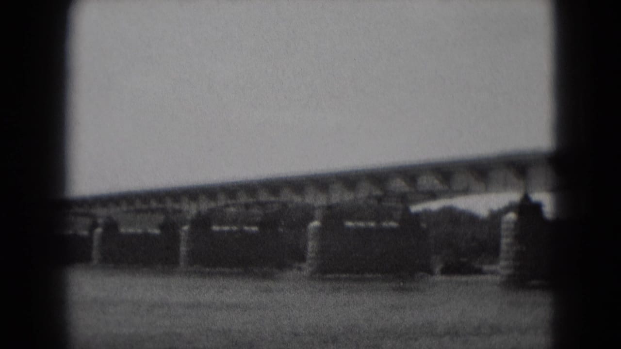 Bridge truss and multiple stone piers stretch across the river under a pale sky.