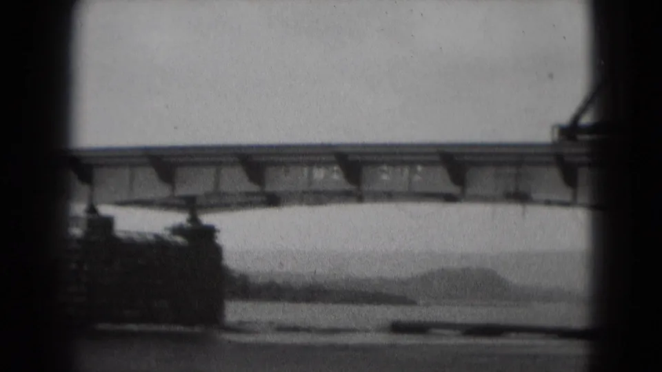 1975 home-movie frame showing the bridge from below, with river water, low hills, and a large support at the left edge.