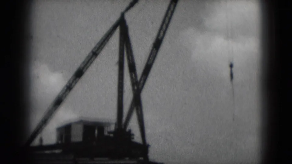 1975 8mm frame of a waterfront crane boom and hanging hook in Middletown, Pennsylvania, with river and sky behind it.