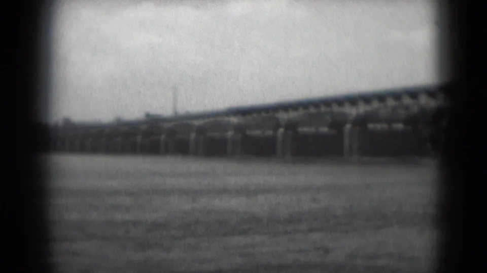 Wide 1975 home-movie view of the bridge over the river near Middletown, with a faint distant vertical structure and soft shoreline detail.